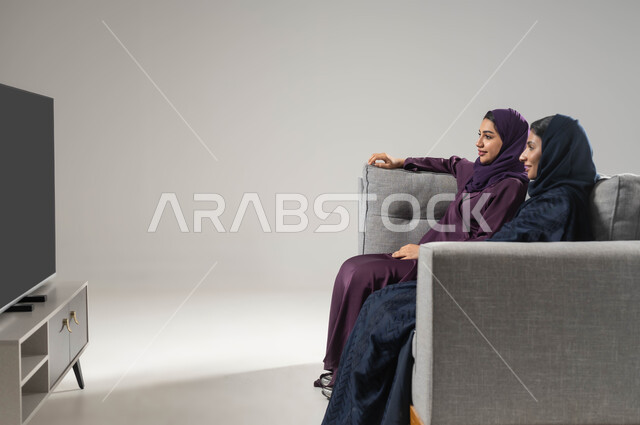 إيماءات وجه و يدين تدل على السعادة والتركيز، بورتريه من الجانب لشابتان عربيتان سعوديتان محجبتان ترتديان العباءة تجلسان على اريكة مريحة، الاستمتاع بقضاء الأوقات الترفيهية العائلية ، مشاهدة الأفلام  المسلسلات مع العائلة، خلفية رمادية