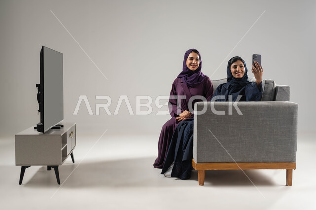النظرإلى كاميرا الموبايل بإيماءات السعادة والسرور، الاستعداد لالتقاط سيلفي، استخدام التكنولوجيا الحديثة، شاشة تلفاز سوداء، بورتريه من الجانب لشابتين عربيتين سعوديتين خليجيتين محجبتان تجلسان على كنبة مريحة تأخدان صور تذكارية، صورة لكامل الجسم، خلفية رمادية