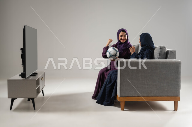 رفع اليدين بتعابير الفوز والانتصار، أنشطة ترفيهية منزلية، متابعة المباريات والالعاب الرياضية، إيماءات الشغف والحماس، بورتريه من الجانب لشابتان عربيتان خليجيتان سعوديتان تجلسان على كنبة تحمل إحداهما كرة، مشاهدة التلفازمع العائلة، صورة لكامل الجسم، خلفية رمادية