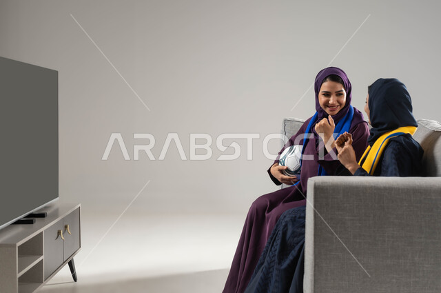 النظر بإيماءات الشغف والحماس، رفع اليدين بتعابير الفوز والانتصار،ارتداء اوشحة تشجيعية، بورتريه لشابتان عربيتان خليجيتان سعوديتان تجلسان على كنبة مريحة تحمل إحداهما كره القدم، متابعة المباريات والالعاب الرياضية، مشاهدة التلفاز مع العائلة، خلفية رمادية