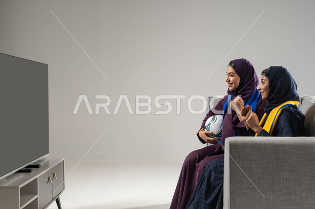 إيماءات الشغف والحماس، رفع اليدين بتعابير الفوز والانتصار،ارتداء اوشحة تشجيعية، بورتريه لشابتان عربيتان خليجيتان سعوديتان تجلسان على كنبة مريحة تحمل إحداهما كره القدم، متابعة المباريات والالعاب الرياضية، مشاهدة التلفاز مع العائلة، خلفية رمادية
