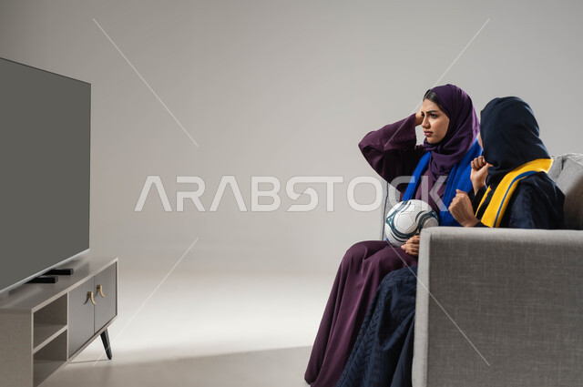 ارتداء اوشحة تشجيعية، بورتريه لشابتان عربيتان خليجيتان سعوديتان تجلسان على كنبة مريحة تحمل إحداهما كره قدم، تعابير الحزن لخسارة الفريق، رفع اليدين بإيماءات الفوز والانتصار، متابعة المباريات و الالعاب الرياضية، مشاهدة التلفاز مع العائلة، خلفية رمادية