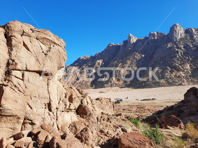 الاستمتاع بالمناظر الطبيعية في وقت النهار، سلسلة الجبال المجاورة لصخرة سبليت في مدينة تبوك الصحراوية، معالم اثرية تاريخية قديمة سياحية مشهورة، نمو النباتات والاعشاب البرية في المناطق المرتفعة، التكوينات والتشكيلات الصخرية والتضاريس في المملكة العربية السعودية