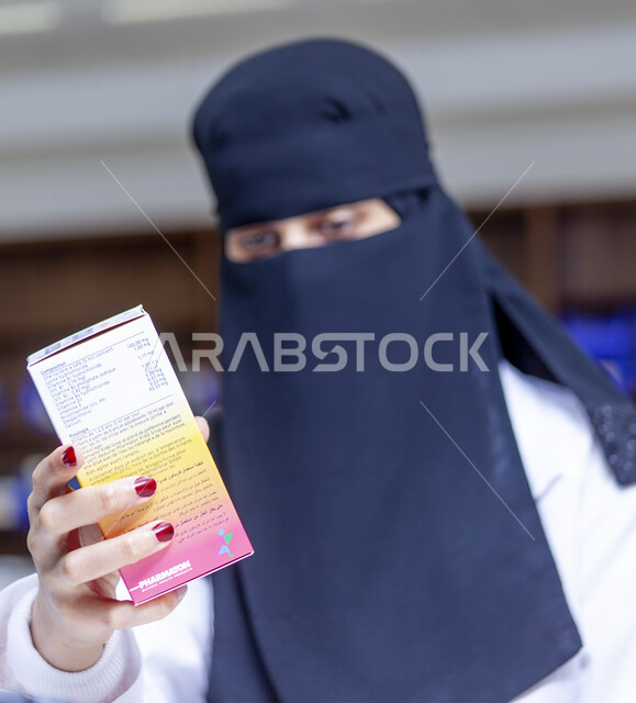 التأكد من صلاحية الأدوية والمستحضرات، صورة مقربة لطبيبة صيدلانية عربية سعودية خليجية منقبة ترتدي المعطف الابيض تمسك بيدها علبة الدواء، تقديم الخدمات العلاجية والاستشارات، خلفية الصيدلية
