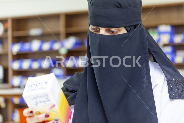 التأكد من صلاحية الأدوية والمستحضرات، صورة مقربة لطبيبة صيدلانية عربية سعودية خليجية منقبة ترتدي المعطف الابيض تمسك بيدها علبة الدواء، تقديم الخدمات العلاجية والاستشارات، خلفية الصيدلية