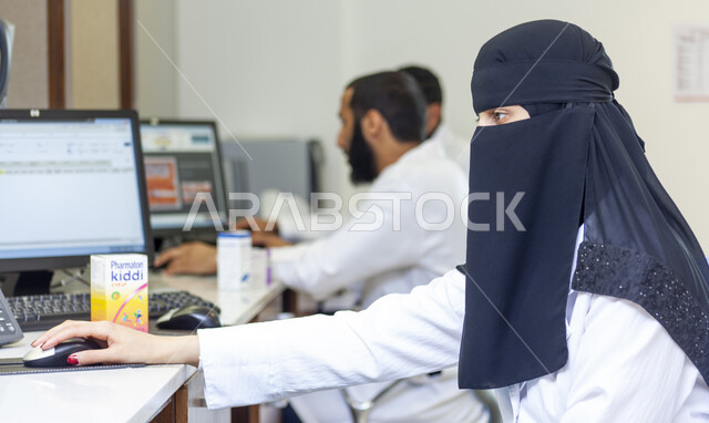 تعاون الزملاء والأطباء داخل صيدلية الجامعة العربية، التأكد من صلاحية الأدوية والمستحضرات، طبيبة صيدلانية عربية سعودية خليجية منقبة ترتدي المعطف الابيض تقوم بالعمل باستخدام جهاز الحاسوب، تقديم الخدمات العلاجية والاستشارات عن بعد
