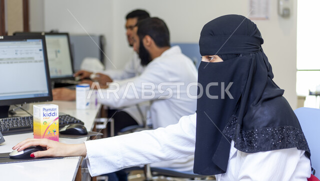 التأكد من صلاحية الأدوية والمستحضرات الطبية، تعاون الزملاء والأطباء داخل صيدلية الجامعة العربية، طبيبة صيدلانية عربية سعودية خليجية منقبة ترتدي المعطف الابيض تقوم بالعمل باستخدام جهاز الحاسوب