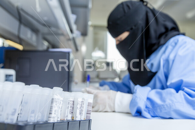 اسخدام أنابيب الاختبار وإجراء التجارب العملية, تطوير التكنولوجيا الحيوية في المختبرات العلمية, صورة مقربة من الجانب لطبيبة مخبرية عربية خليجية سعودية ترتدي المعطف الطبي والقفازات تقوم بتدوين وتسجيل نتائج التحليل الكيميائي, العمل في مخابر المملكة العربية السعودية