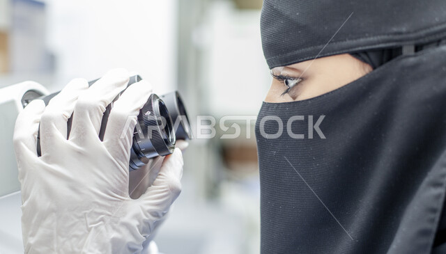 مهن ووظائف نسائية, أجهزة الفحص الدقيقة للمواد الكيمائية, صورة مقربة من الجانب لطبيبة مخبرية عربية خليجية سعودية منقبة ترتدي المعطف الطبي والقفازات تقوم بفحص العينات عبر جهاز المجهر في مخبر الأبحاث الطبية المتطور, تطور التكنولوجيا الحيوية في المختبرات العلمية