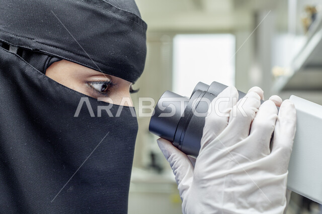 Women's professions and jobs, accurate testing devices for chemical substances, a close-up picture from the side of a veiled Saudi Arab Gulf laboratory doctor wearing a medical coat and gloves examining samples through a microscope in an advanced medical research laboratory, the development of biotechnology in scientific laboratories