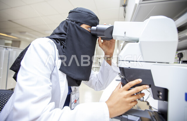 أجهزة الفحص الدقيقة للمواد الكيمائية, صورة مقربة من الجانب لطبيبة مخبرية عربية خليجية سعودية منقبة ترتدي المعطف الطبي الأبيض تقوم بفحص العينات عبر جهاز المجهر في مخبر الأبحاث الطبية المتطور, تطور التكنولوجيا الحيوية في المختبرات العلمية, مهن ووظائف نسائية