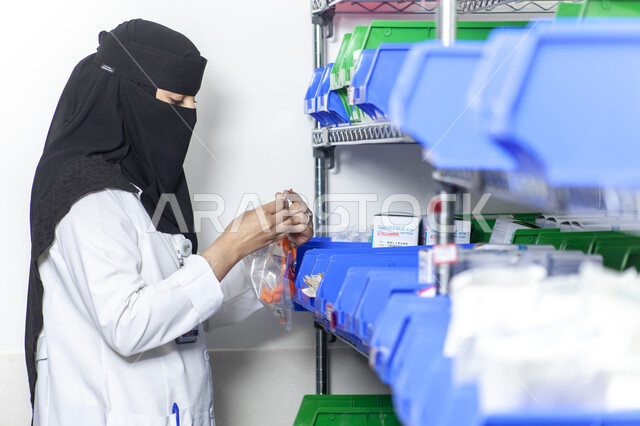 إجراء التجارب العملية, صورة مقربة من الجانب لطبيبة مخبرية عربي خليجية سعودية منقبة ترتدي المعطف الطبي الأبيض تمسك مواد التحليل المخبري, تطور التكنولوجيا الحيوية في المختبرات العلمية, تطوير مهارات البحث الطبي في معامل الكيمياء في المملكة العربية السعودية