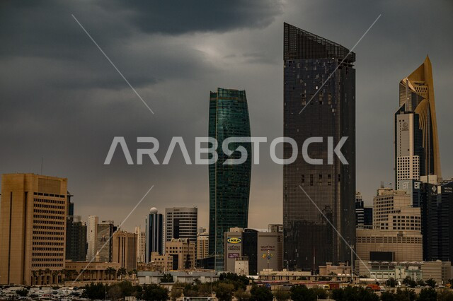 The artistic architecture of the Kuwait City towers during the day, the view of the sky filled with dark clouds, the development and urban progress of the Kuwaiti capital’s facilities, famous tourist attractions, trees and green plants on the city’s roads