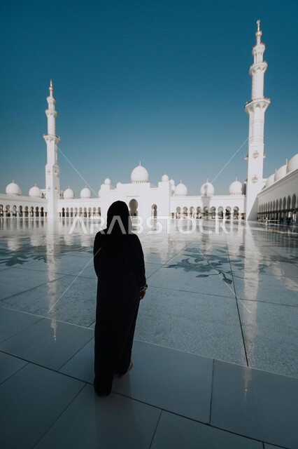 Visiting religious landmarks in the United Arab Emirates, worship and getting closer to God, a picture from behind of an Arab woman walking around the outer courtyard of the Sheikh Zayed bin Sultan Al Nahyan Mosque in Abu Dhabi, architectural engineering art and designs in the Islamic style in mosques, Islamic background