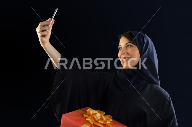 بورتريه مرأه سعودية خليجية تحمل صندوق هدايا كبير ، تستخدم هاتفها المحمول لتصوير صورة سلفي  ، شراء الهدايا ،  الترفيه و التسوق ، يوم التسوق ، الجمعة البيضاء