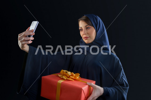 Portrait of a Saudi Arabian Gulf woman carrying a large gift box, using her mobile phone to photograph a selfie, buying gifts, entertainment and shopping, shopping day, White Friday