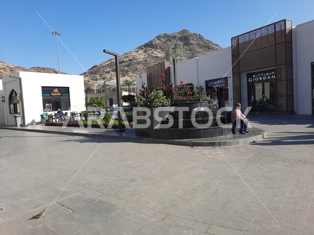 الفن الهندسي المعماري على الطراز الحديث في مكة المكرمة، رجل يجلس على حافة الحوض المزروع بالورود بالقرب من المحلات التجارية في مركز حراء الثقافي، اقتناء الهدايا والتذكارات، أماكن إسلامية مقدسة، معالم سياحية مشهورة في المملكة العربية السعودية
