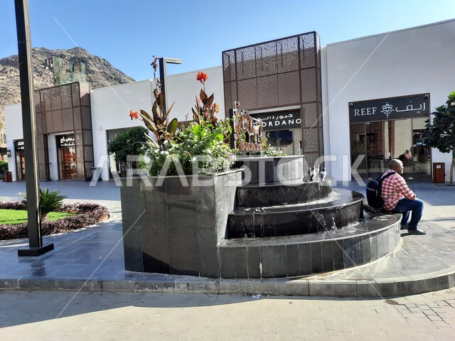 الفن الهندسي المعماري على الطراز الحديث في مكة المكرمة، رجل يجلس على حافة الحوض المزروع بالورود بالقرب من المحلات التجارية في مركز حراء الثقافي، اقتناء الهدايا والتذكارات، أماكن إسلامية مقدسة، معالم سياحية مشهورة في المملكة العربية السعودية