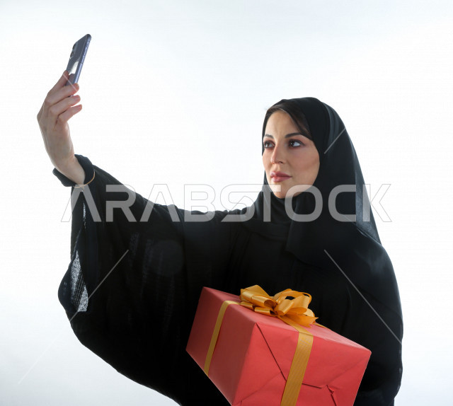 بورتريه مرأه سعودية خليجية تحمل صندوق هدايا كبير ، تستخدم هاتفها المحمول لتصوير صورة سلفي  ، شراء الهدايا ،  الترفيه و التسوق ، يوم التسوق ، الجمعة البيضاء