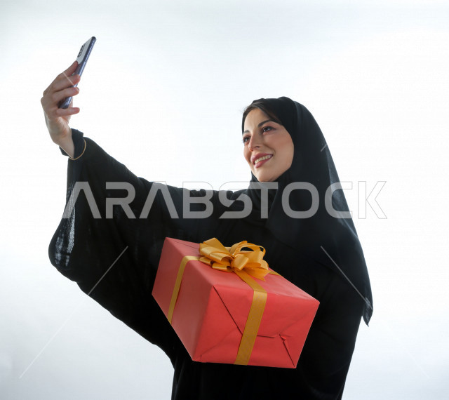 بورتريه مرأه سعودية خليجية تحمل صندوق هدايا كبير ، تستخدم هاتفها المحمول لتصوير صورة سلفي  ، شراء الهدايا ،  الترفيه و التسوق ، يوم التسوق ، الجمعة البيضاء