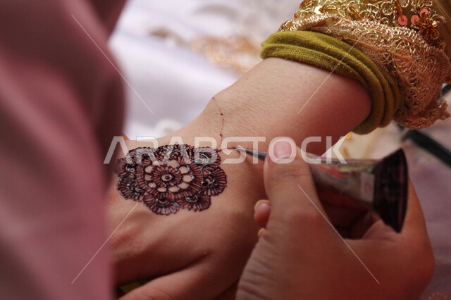  زخارف وفنون يدويه من مواد طبيعيه، التجمل والتزيين والاهتمام بالمظهر الخارجي، عادات وتقاليد في المناسبات والأفراح السعيدة، صورة مقربة ليد إمرأة عربية خليجية سعودية تقوم بالرسم والنقش على يد فتاة باستخدام الحناء الحمراء، مهن تقليدية شعبية نسائية