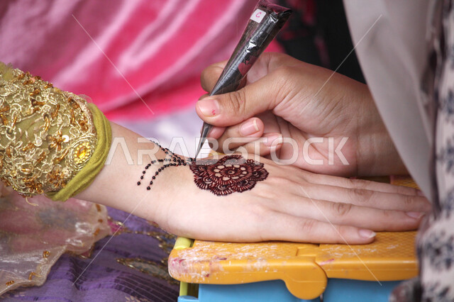 التجمل والتزيين والاهتمام بالمظهر الخارجي، عادات وتقاليد في المناسبات والأفراح السعيدة، صورة مقربة ليد إمرأة عربية خليجية سعودية تقوم بالرسم والنقش على يد فتاة باستخدام الحناء الحمراء، مهن تقليدية شعبية نسائية، زخارف وفنون يدويه من مواد طبيعيه