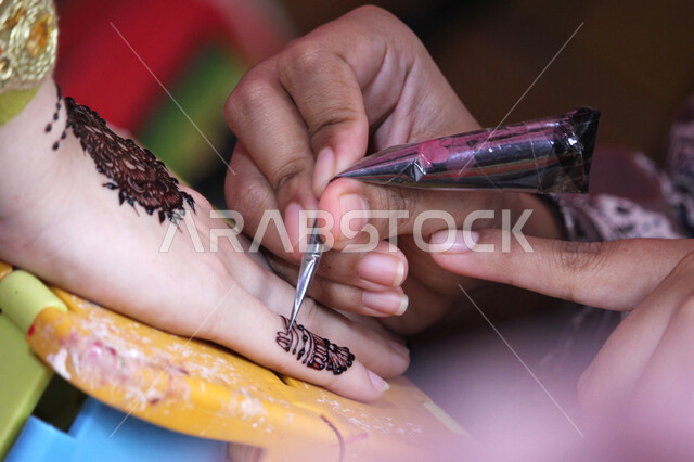  زخارف وفنون يدويه من مواد طبيعيه، التجمل والتزيين والاهتمام بالمظهر الخارجي، عادات وتقاليد في المناسبات والأفراح السعيدة، صورة مقربة ليد إمرأة عربية خليجية سعودية تقوم بالرسم والنقش على يد فتاة باستخدام الحناء الحمراء، مهن تقليدية شعبية نسائية