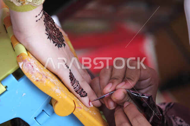 عادات وتقاليد في المناسبات والأفراح السعيدة، صورة مقربة ليد إمرأة عربية خليجية سعودية تقوم بالرسم والنقش على يد فتاة باستخدام الحناء الحمراء، التزين والتجمل والاهتمام بالمظهر الخارجي، مهن تقليدية شعبية نسائية، زخارف وفنون يدويه من مواد طبيعيه