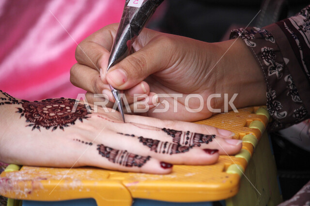 التزين والتجمل والاهتمام بالمظهر الخارجي، عادات وتقاليد في المناسبات والأفراح السعيدة، صورة مقربة ليد إمرأة عربية خليجية سعودية تقوم بالرسم والنقش على يد فتاة باستخدام الحناء الحمراء، مهن تقليدية شعبية نسائية، زخارف وفنون يدويه من مواد طبيعيه