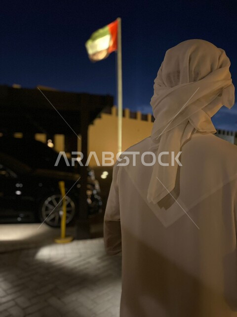 إحياء ذكرى الاستقلال لدولة الإمارات العربية المتحدة، صورة من الخلف لرجل عربي خليجي إماراتي يرتدي العمامة والكندورة الإماراتية، الاحتفال بالعيد الوطني الإماراتي، سارية علم الوطن ترفرف في الهواء الطلق في إمارة الشارقة