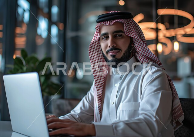مهن ووظائف مكتبية, صورة مقربة لرجل عربي خليجي سعودي يرتدي الشماغ والثوب التقليدي ينظر إلى الكاميرا يقوم باستخدام الحاسب المحمول, إدارة وتنظيم الأعمال ضمن مقر الشركة، دمج التقنية والتكنولوجيا مع الحياة العملية، بيئة العمل المريحة في الشركات السعودية