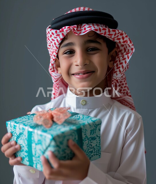 الشعور بالسعادة والفرح, بورتريه مقرب لفتى عربي خليجي سعودي مبتسم يرتدي الشماغ والثوب التقليدي ينظر إلى الكاميرا يحمل بيديه علبة هدية مغلفة، مناسبات وأعياد سعيدة، تقديم الهدايا والتهنئات, خلفية رمادية