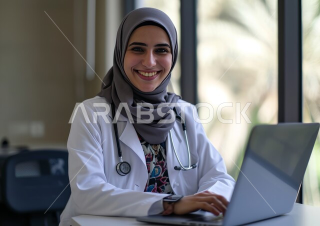 Using modern technologies in Saudi medical centers and hospitals, providing medical consultations and therapeutic services to patients via the Internet, a smiling Saudi Arabian Gulf doctor wearing a white medical coat and an examination stethoscope looking at the camera and using a laptop