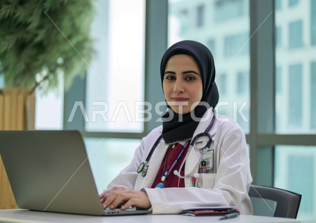 تقديم الخدمات العلاجية في المراكز الطبية والمستشفيات في السعودية, النظر إلى الكاميرا بابتسامة, استخدام الأجهزة التقنية الحديثة في المجال الطبي, صورة مقربة لطبيبة عربية خليجية سعودية محجبة ترتدي المعطف الطبي الأبيض وسماعة الفحص تستخدم الحاسب المحمول