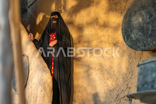 Commemorating the founding day of the first Saudi state, February 22, a Saudi Gulf Arab woman wearing a burqa and black abaya stands in an old-style mud heritage house, a day in the year 1727 AD, the pride and honor of belonging to the homeland, wearing popular costumes on national occasions and holidays.