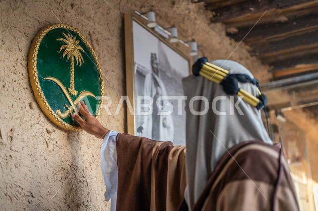 ذكرى تأسيس الدولة السعودية الاولى 22 فبراير، يوم بدينا 1727 م، ارتداء الأزياء الشعبيه للأعياد و المناسبات الوطنية، صورة مقربة من الخلف لرجل عربي خليجي سعودي يرتدي الزي التقليدي والعقال يقوم بلمس شعار النخلة والسيفين في أحد البيوت الطينية التراثيه القديمة