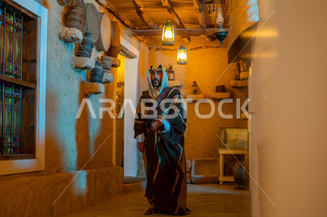 Wearing popular costumes for national occasions and holidays, commemorating the founding of the first Saudi state on February 22, showing love, loyalty and belonging to the homeland, Day of Dina 1727 AD, a Saudi Gulf Arab man wearing a traditional costume and headband walking around one of the old traditional mud houses looking at something