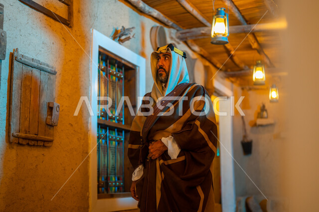 Anniversary of the founding of the first Saudi state, February 22, showing love, loyalty, and belonging to the homeland, wearing popular costumes for national occasions and holidays, Day of Dina 1727 AD, a Saudi Gulf Arab man wearing traditional clothing and a headband standing in one of the old traditional mud houses looking at something