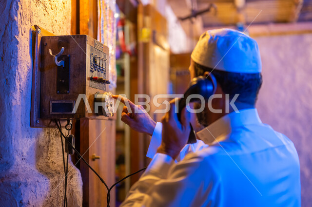 Antiques and old antique devices, traditional means of communication in Saudi Arabia, the anniversary of the founding of the first Saudi state, February 22, a close-up photo from the back of a Saudi Gulf Arab man wearing a traditional dress and a white hat, using the landline phone in one of the traditional markets, Day of Dina 1727 AD.