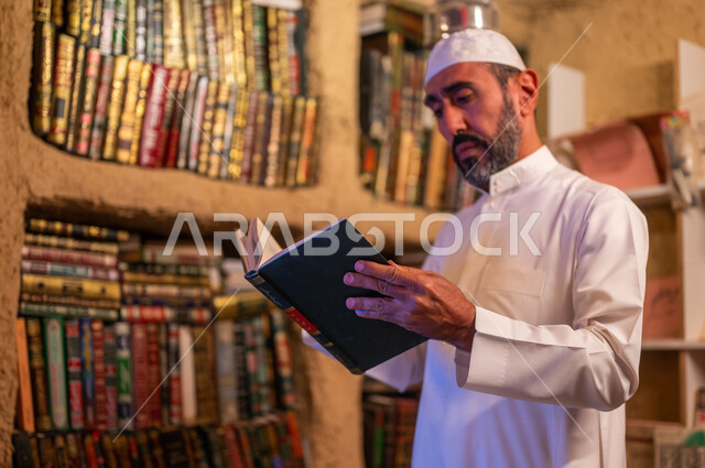 The concept of culture and self-learning, supporting and developing the linguistic and scientific repertoire, a Saudi Gulf Arab man wearing a traditional dress and a white hat reading a book in the library built in the old style, the anniversary of the founding of the first Saudi state, February 22, the day of our religion 1727 AD.