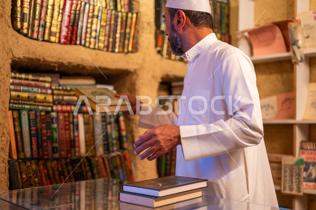 دعم وتنمية الذخيرة اللغوية والعلمية, مكان القراءة ومحبي العلم والمعرفة، مفهوم الثقافة والتعلم الذاتي, رجل عربي خليجي سعودي يرتدي الثوب التقليدي والقبعه البيضاء يقوم بترتيب الكتب على الرفوف في المكتبة, ذكرى تأسيس الدولة السعودية الأولى 22 فبراير، يوم بدينا 1727 م