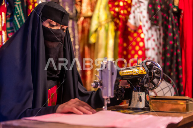 أعمال وحرف ومهن تقليدية نسائية, ذكرى تأسيس الدوله السعوديه الاولى ٢٢ فبراير ،يوم التأسيس 1727 م، صورة مقربة لخياطة عربية سعودية خليجية ترتدي العباءة السوداء والبرقع تقوم بحياكة وخياطة القماش على الماكينة اليدوية في الأسواق الشعبيه القديمة في السعودية