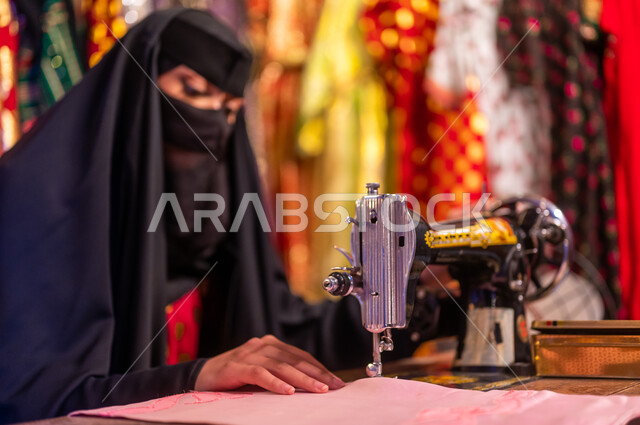ذكرى تأسيس الدوله السعوديه الاولى ٢٢ فبراير ،يوم التأسيس 1727 م، أعمال وحرف ومهن تقليدية نسائية, صورة مقربة لخياطة عربية سعودية خليجية ترتدي العباءة السوداء والبرقع تقوم بحياكة وخياطة القماش على الماكينة اليدوية في الأسواق الشعبيه القديمة في السعودية