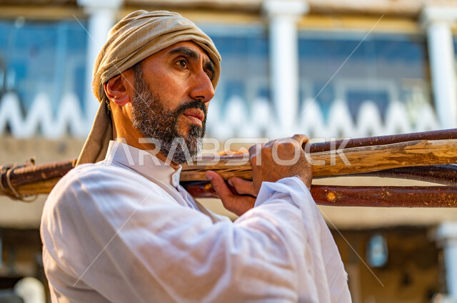 Traditional Bedouin lifestyle, ancient heating methods, Day of Bedina 1727 AD, anniversary of the founding of the first Saudi state, February 22, collecting firewood in the winter season, a close-up photo of a Saudi Arabian Gulf farmer wearing a traditional dress carrying a group of heavy wood sticks on his shoulder inside traditional houses
