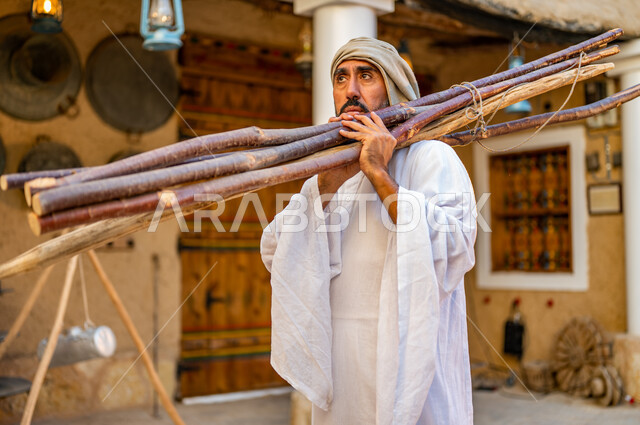 ذكرى تأسيس الدولة السعودية الأولى 22 فبراير، تجميع الحطب في موسم الشتاء, فلاح عربي خليجي سعودي يرتدي الزي الشعبي يحمل مجموعه من أعواد الخشب الثقيلة على كتفه داخل البيوت التراثية، يوم بدينا 1727 م، الحياة التقليدية البدوية، وسائل التدفئة القديمة