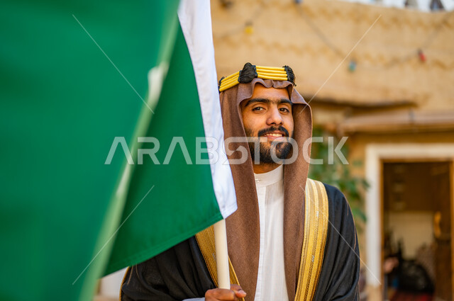 الفخر والاعتزاز بإنجازات الوطن، شعار الهوية الوطنية وراية الإسلام والتوحيد، يوم بدينا 1727 م، صورة مقربة لشاب عربي خليجي سعودي مبتسم يرتدي العقال الذهبي والبشت يحمل بيده علم التأسيس في بيت طيني تراثي, ذكرى تأسيس الدولة السعودية الأولى 22 فبراير 