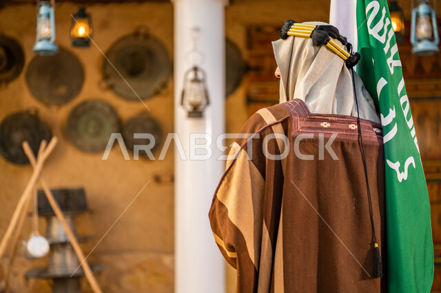 ذكرى تأسيس الدولة السعودية الأولى 1727 م، يوم بدينا 22 فبراير، صورة من الخلف لرجل عربي خليجي سعودي يرتدي العقال الذهبي والبشت يحمل بيده علم التأسيس في بيت طيني تراثي, الاعتزاز والفخر بإنجازات الوطن، شعار الهوية الوطنية وراية الإسلام والتوحيد