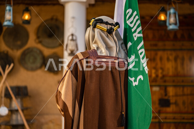 The emblem of national identity and the banner of Islam and monotheism, the anniversary of the founding of the first Saudi state in 1727 AD, the day of our religion, February 22, a picture from the back of a Saudi Gulf Arab man wearing a golden iqal and a bisht, holding in his hand the founding flag in a heritage mud house, pride in the nation’s achievements.