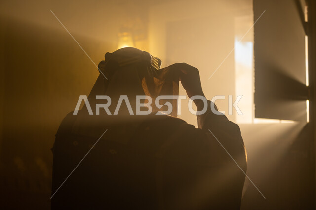 الأناقة والاهتمام بالمظهر الخارجي, مفهوم الشموخ والاعتزاز والفخر, الأعياد والمناسبات الوطنية, ذكرى تأسيس الدولة السعودية الأولى 22 فبراير, صورة مقربة من الخلف لرجل عربي خليجي سعودي يرتدي البشت والعقال يقوم بتعديل لباسه يقف بجانب النافذة, يوم بدينا 1727 م