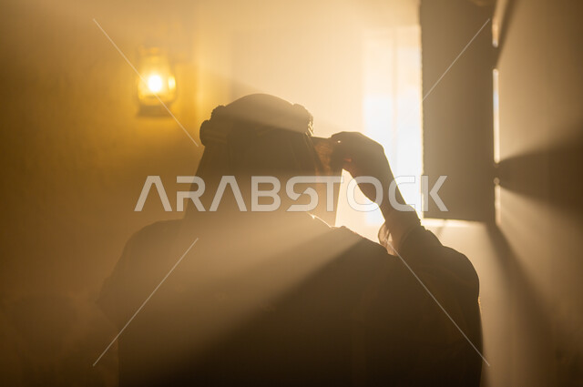 الاهتمام بالأناقة والمظهر الخارجي, مفهوم الشموخ والاعتزاز والفخر, الأعياد والمناسبات الوطنية, ذكرى تأسيس الدولة السعودية الأولى 22 فبراير, صورة مقربة من الخلف لرجل عربي خليجي سعودي يرتدي البشت والعقال يقوم بتعديل لباسه يقف بجانب النافذة, يوم بدينا 1727 م